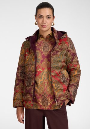 Frau trägt eine bunte Steppjacke mit Paisley-Muster über einem passenden Hemd, kombiniert mit dunkelbrauner Hose, steht vor weißem Hintergrund.