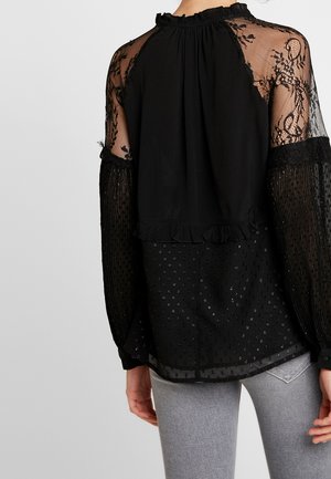 Person iført en sort gennemsigtig blondebluse med mønstrede lange ærmer og lysegrå jeans, vist fra ryggen mod en ensfarvet baggrund.