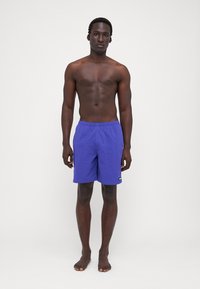 Purple swim shorts with an elastic waistband, a small logo patch, and a lightweight fabric. Model stands barefoot against a neutral background.