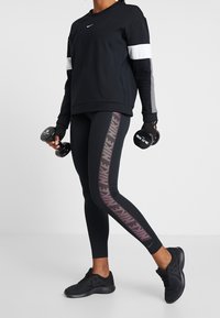 Czarna sportowa bluza z szarymi i białymi akcentami, w połączeniu z czarnymi legginsami z różowym wzorem w paski, trzymająca hantle.
