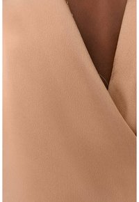 Beige slyngebluse med en glat, tekstureret stof og en dyb V-udskæring. Stoffet falder blødt og fremhæver dens elegante silhuet.