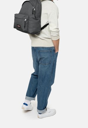Personne portant un pull beige clair, un jean bleu avec revers, des baskets blanches, et portant un grand sac à dos gris foncé sur une épaule.