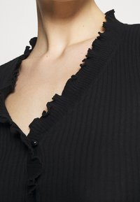 Svart ribbad cardigan med volangkrage och svarta knappar som accent. Tygets struktur är mjuk och något stretchig.