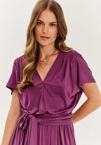 Fialové šaty s výstrihom do V, vyrobené z jemnej tkaniny, so skrátenými rukávmi, opaskom v páse a splývavou sukňou s prešívaniami pre dodatočnú textúru.