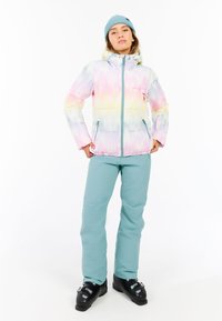 Geplüschte Jacke im pastellfarbenen Batikmuster mit einem Reißverschluss, zwei seitlichen Taschen und einer Kapuze, kombiniert mit hellblauen Hosen und schwarzen Stiefeln.