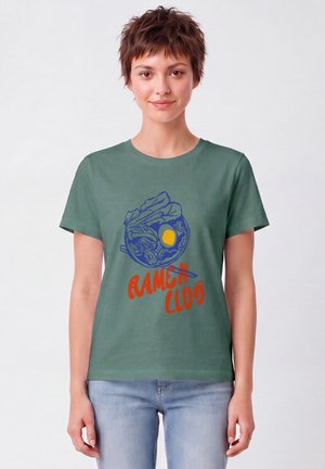 Jeune personne aux cheveux courts portant un T-shirt vert avec un bol de ramen et le texte "Ramen Club,", assorti à un jean bleu clair, debout devant un fond uni.