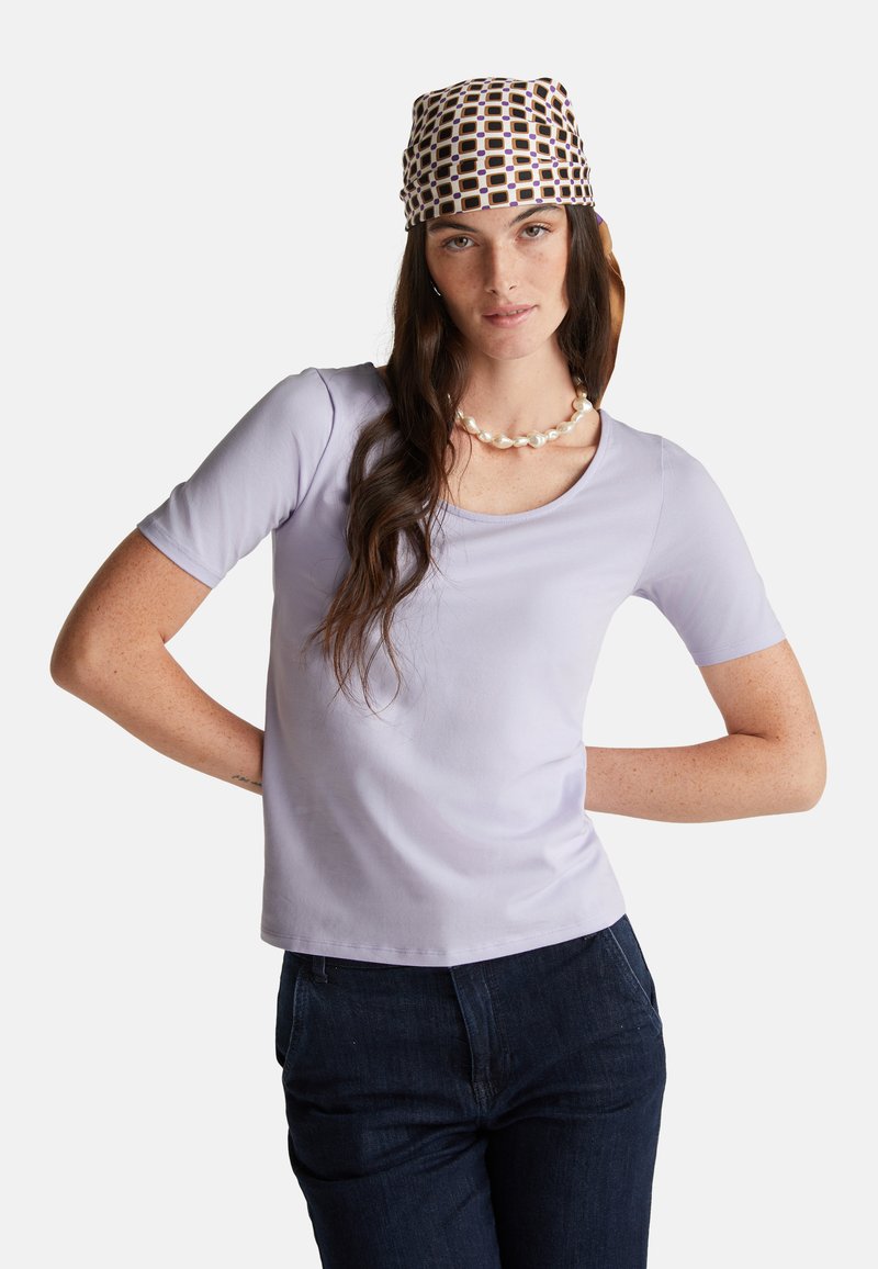 Camicia corta a maniche corte di color lavanda chiaro con scollatura a barca, abbinata a pantaloni in denim scuro. Accentuata da un foulard stampato e una collana di perle.