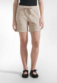 Kobieta ubrana w beżowe szorty z podwiniętymi brzegami, z przednimi kieszeniami i sznurkiem w talii, zestawione z czarnymi, sznurowanymi butami na płaskiej podeszwie.