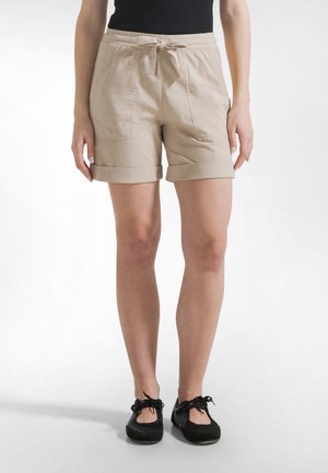 Kobieta ubrana w beżowe szorty z podwiniętymi brzegami, z przednimi kieszeniami i sznurkiem w talii, zestawione z czarnymi, sznurowanymi butami na płaskiej podeszwie.
