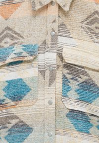 Chemise en coton à boutons avec un motif géométrique beige, bleu et orange. Dotée de boutons pression et de deux poches avant. Tissu texturé.