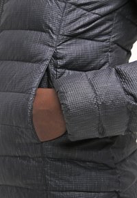 Mörkgrå, texturerad puffjacka med en hand inskjuten i en sidoficka, som visar quiltat sömnad och polstrade tygdetaljer.