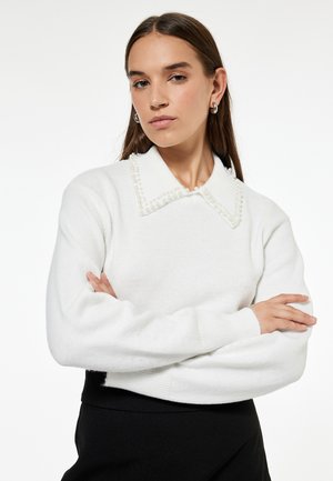 Junge Frau mit langen braunen Haaren trägt einen weißen Pullover mit perlenverziertem Kragen und silbernen Creolen, verschränkt die Arme.