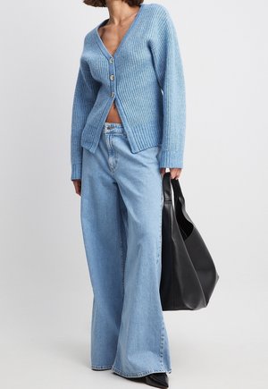 Femme portant un cardigan bleu clair boutonné et un jean à jambes larges, tenant un grand sac cabas en cuir noir sur un fond uni.