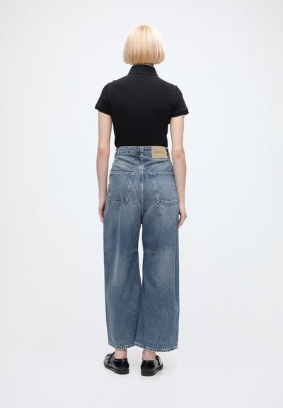 Personne aux cheveux blonds courts portant un haut noir à manches courtes, un jean bleu taille haute à jambes larges et des chaussures noires, face à l'arrière de la caméra.