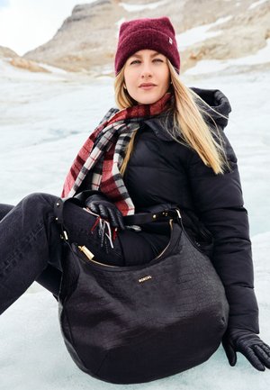 Sacs en cuir noir texturé avec matériel doré, associé à une écharpe à carreaux rouge et noir, une veste matelassée noire, des gants et un fond enneigé.