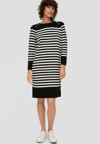 Gestreiftes Strickkleid in schwarz und weiß mit rundem Halsausschnitt, Schulterknöpfen, langen Ärmeln und schwarzem Saum, kombiniert mit weißen Sneakern.