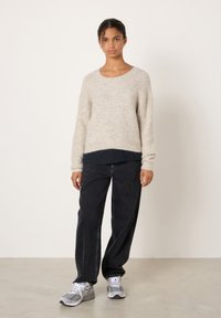 Jauna sieviete stāv, valkājot bēša trikotāžas džemperi, melnas vaļīgas bikses, pelēkas un baltas sporta kurpes, pret vienkāršu gaišu fonu.