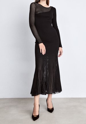 Femme portant une robe noire à manches longues côtelée avec des détails rayés transparents sur les manches et le bas de la jupe, associée à des escarpins noirs à bouts pointus.