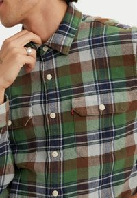 Camisa de franela a cuadros verdes y marrones con cierre de botones, dos bolsillos en el pecho y una sutil etiqueta del logo de Levi's en el bolsillo izquierdo.