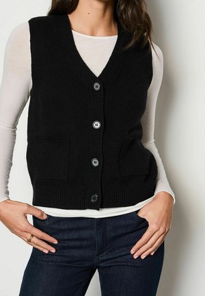 Vrouw draagt een zwart gebreid vest met knopen over een wit shirt met lange mouwen en donkere jeans, handen rustend op de heupen in een neutrale houding.