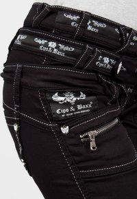 Zwarte jeans met witte stiksels, meerdere zakken en merknamen. Inclusief een ritszak en decoratieve hardware.