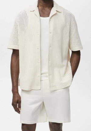 Homme portant une chemise en maille couleur crème à manches courtes par-dessus un débardeur blanc, avec un short assorti couleur crème.