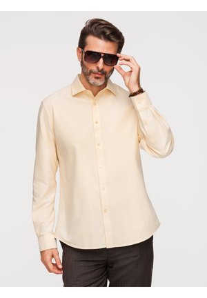 Mand med trimmet skæg iført beige skjorte og mørke solbriller, justerer briller med højre hånd mod ensfarvet baggrund.