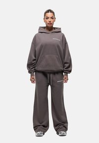 Felpa marrone oversize con tasca frontale e logo, abbinata a pantaloni della tuta a gamba larga dello stesso colore e sneakers camouflage.