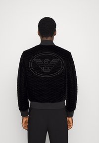 Svart kviltad bomberjacka med ribbad mudd och nederkant. Har en stor präglad örnlogga på baksidan. Mjuk textur.