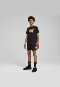 Camiseta de algodón negra con logo "PUMA" naranja, combinada con pantalones cortos negros con acento de logo. Zapatillas y calcetines negros completan el look.
