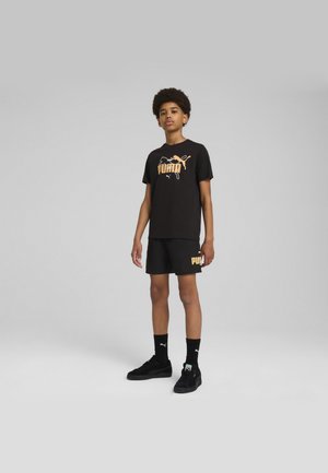 Camiseta de algodón negra con logo "PUMA" naranja, combinada con pantalones cortos negros con acento de logo. Zapatillas y calcetines negros completan el look.