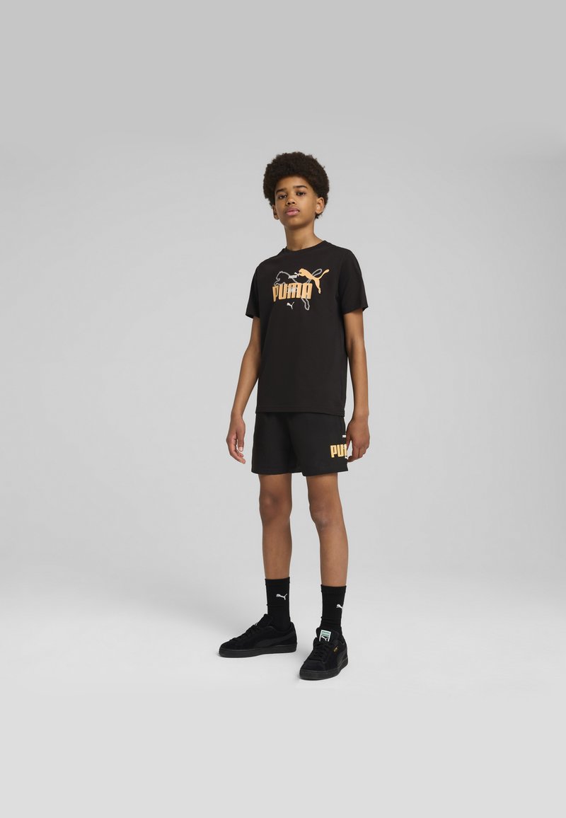 Camiseta de algodón negra con logo "PUMA" naranja, combinada con pantalones cortos negros con acento de logo. Zapatillas y calcetines negros completan el look.