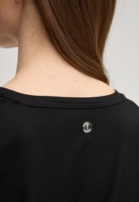 T-shirt in cotone nero con scollo ampio, caratterizzata da un piccolo logo rotondo argento sul retro vicino al collo. Texture liscia.