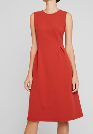 Femme portant une robe rouge sans manches, longueur genoux, coupe évasée, sur un fond gris clair uni.