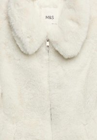 Vit fuskpälsjacka med stor krage, dragkedja fram och subtila texturdetaljer. Märkesetikett "M&S" fäst nära halslinningen.