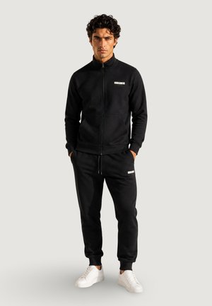 Mand stående iført en sort lynlåstrøje og sorte joggers med hvide sneakers på en ensfarvet lys baggrund.