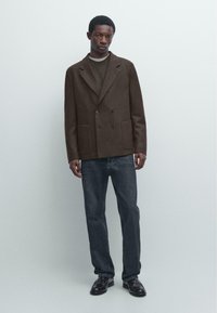 Brązowy dwurzędowy płaszcz z dużymi przednimi kieszeniami, w połączeniu z ciemnymi dżinsami i czarnymi butami. Płaszcz ma kołnierz z wycięciem i gładką fakturę.