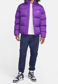 Osoba oblečená do fialového puffer kabátu Nike, bílého trička, námořnických cargo kalhot a bílých s námořnickými akcenty vysokých tenisek Nike, stojící na bílém pozadí.