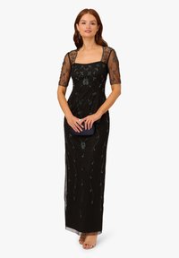 Schwarzes Abendkleid mit quadratischem Ausschnitt, verziert mit Perlenstickerei in gewundenen Mustern und durchsichtigen, mit Perlen besetzten Ärmeln. Begleitet von einer Clutch.