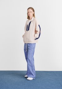 Casaco de vento rosa claro com detalhes em azul-marinho e design de meia-zip, combinado com calças azul-claro com costura branca. Tecido texturizado.