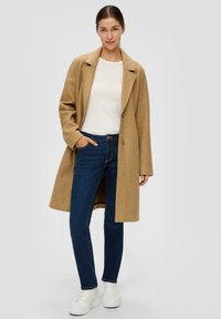 Long manteau en laine couleur camel avec rabats et deux boutons, associé à un jean bleu sombre et des baskets blanches à semelle épaisse.