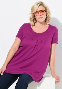 Top a maniche corte, dalla vestibilità ampia, in tessuto viola vivace con scollatura a barca e piccole pieghe nella parte anteriore, abbinato a pantaloni scuri.