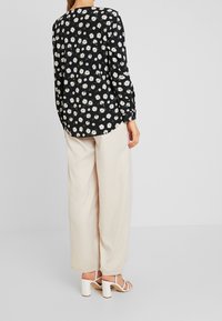 Blouse noire à fleurs avec des marguerites blanches, manches longues et ourlet arrondi, associée à un pantalon large crème et des sandales blanches à talons.