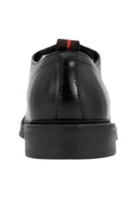 Scarpe in pelle nera con finitura liscia, tacco basso e una linguetta di accento rossa e nera sul tallone. Dettagli cuciti lungo le cuciture.