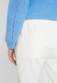 Maglione blu lavorato a maglia con motivo testurizzato, abbinato a pantaloni bianchi con una tasca posteriore visibile e vita elastica.