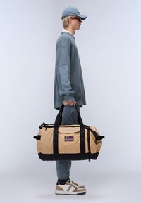 Beige duffel bag with black accents, featuring a Norwegian flag patch, sturdy handles, and a zipper closure. Worn by person in casual attire.