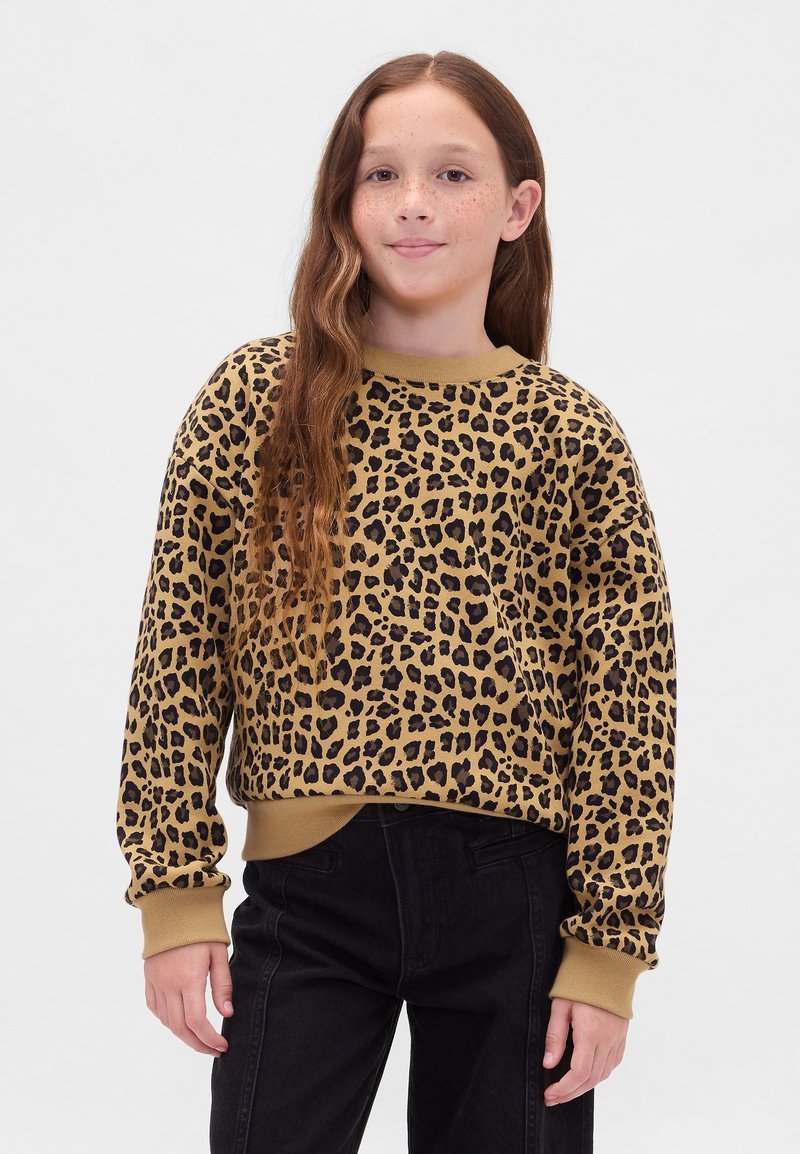 Niña con cabello largo y ondulado, vistiendo un suéter marrón y negro con estampado de leopardo y pantalones negros, de pie contra un fondo claro y sencillo.