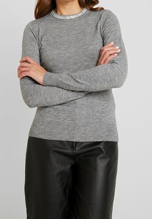 Frau trägt einen grauen Langarm-Pullover mit Strasssteinkragen und schwarze Lederhose, verschränkte Arme vor einem schlichten Hintergrund.
