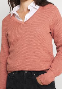 Rosa Strickpullover mit V-Ausschnitt und strukturiertem Muster. Lange Ärmel mit gerippten Bündchen. Über einem weißen Hemd mit Kragen getragen.