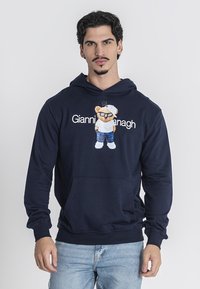 Námořnická modrá mikina vyrobená z měkké látky, s grafikou medvěda v slunečních brýlích, pod kterou je vytištěn text "Gianni Hanagh". Elastické manžety.